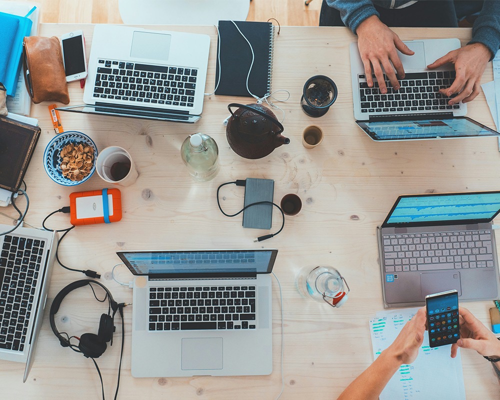 Overhead view of a wooden table with laptops, gadgets, and drinks.