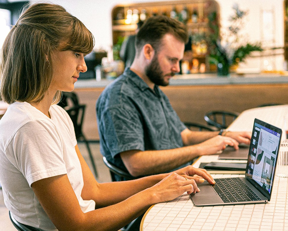 Two people working on laptops at a table inside a community area.